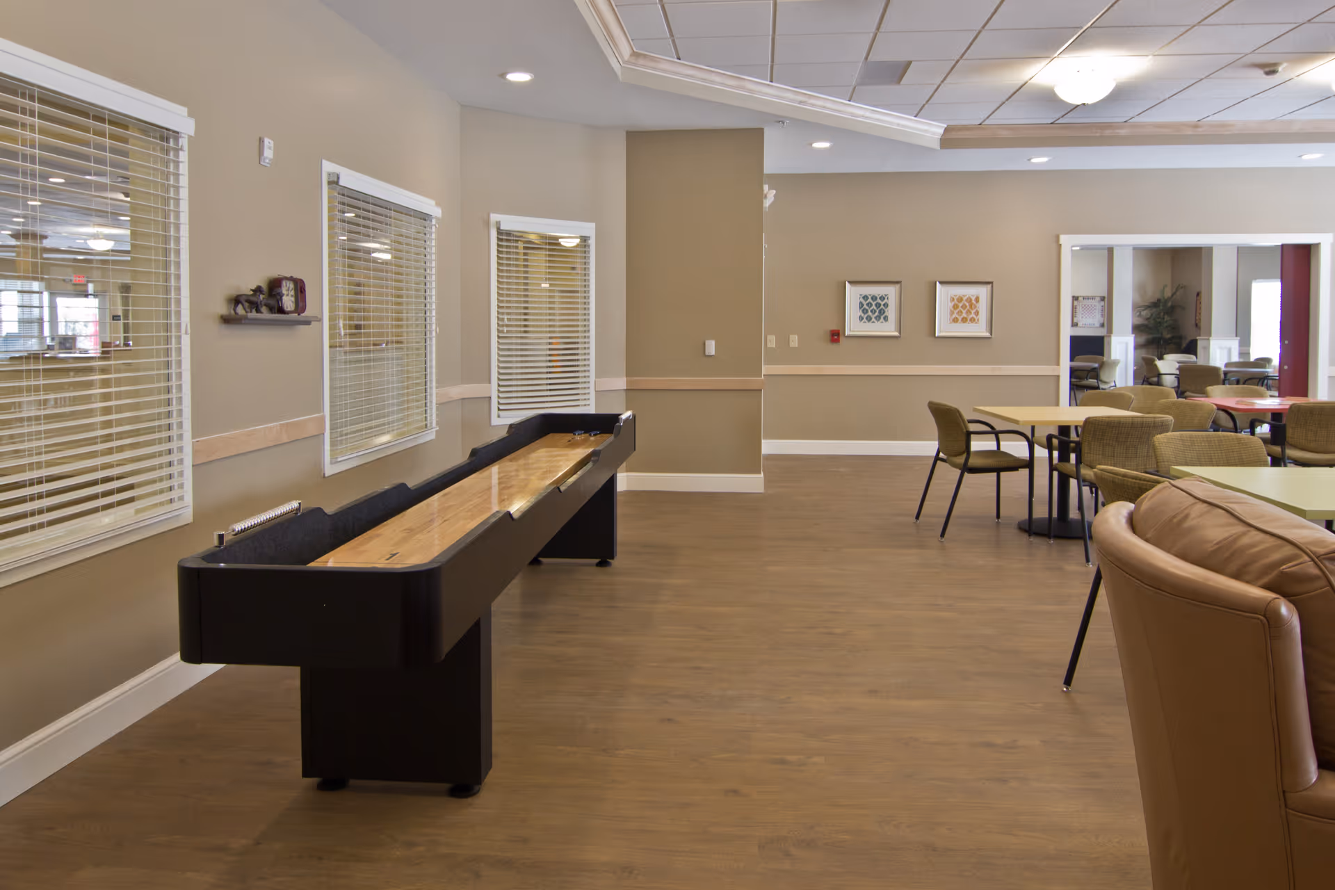 A spacious common area in a senior living facility featuring a shuffleboard table along the left wall, several tables and chairs arranged for socializing or dining, beige walls with white trim, and a brown leather armchair in the foreground.
