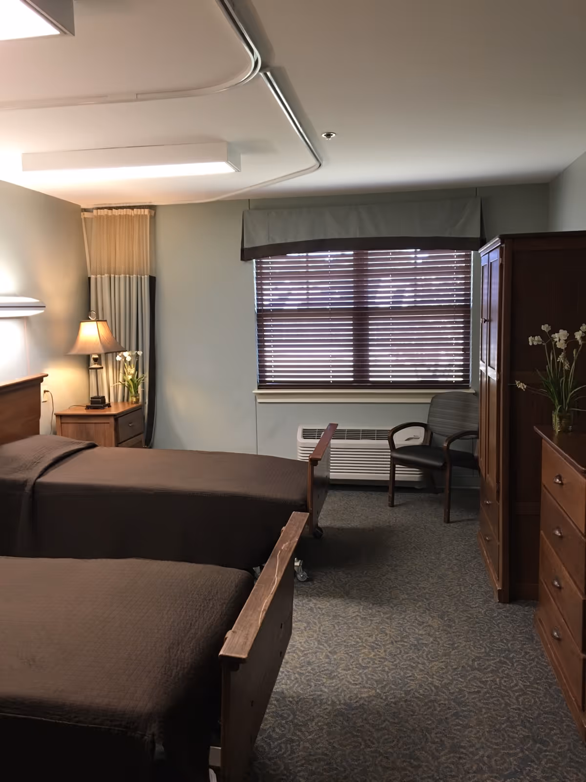 Two single beds in a senior living bedroom with a nightstand and lamp, wooden furniture, and a window with closed blinds.