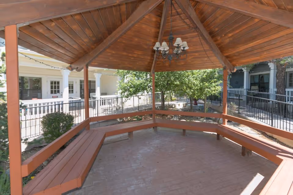 A wooden gazebo with a pitched roof and a chandelier hanging from the center. The gazebo has built-in wooden benches along the perimeter and is situated outdoors with trees and a building with white columns and railings visible in the background.