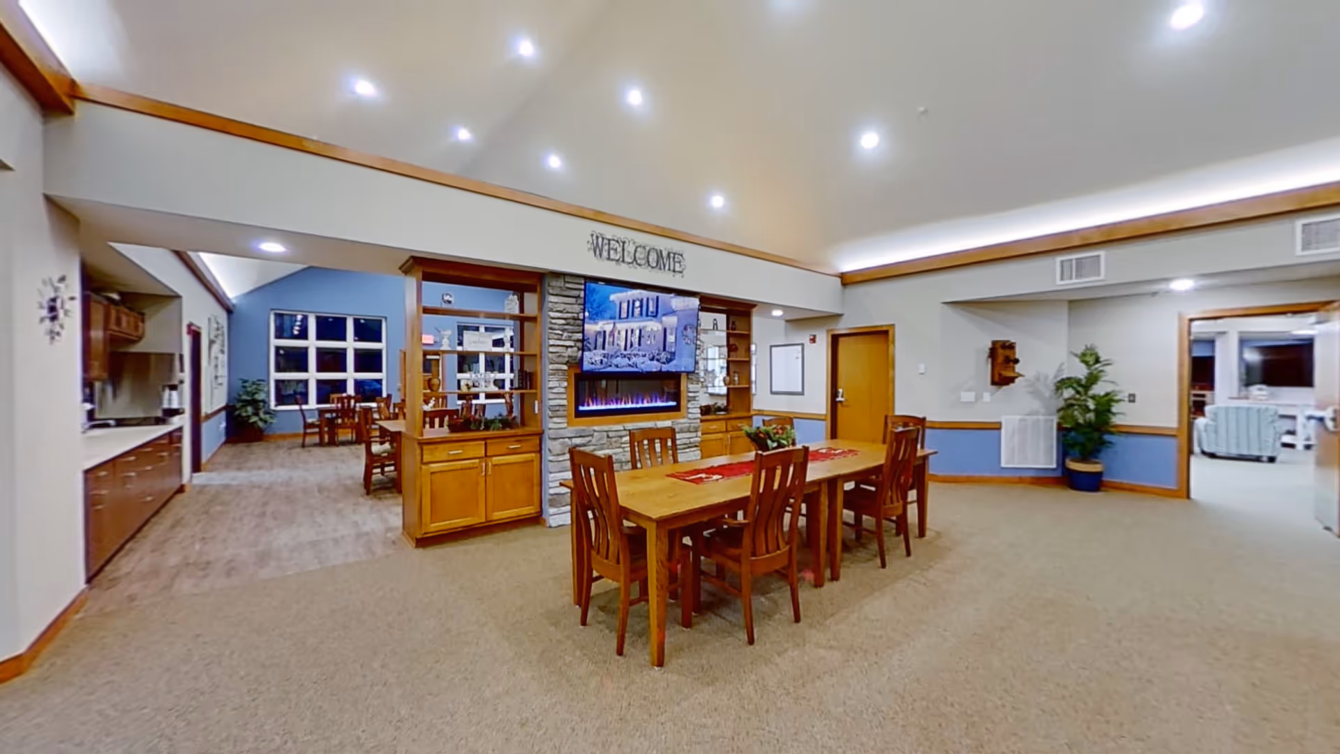 Interior view of a senior living facility common area with a wooden dining table and six chairs in the center. Behind the table is a stone fireplace with a mounted TV above it and wooden cabinets on either side. The walls are painted in neutral tones with blue accents, and the ceiling has recessed lighting. To the left, there is a kitchen area with cabinets and appliances, and in the background, a dining area with additional tables and chairs. To the right, there is a doorway leading to a living room with seating.