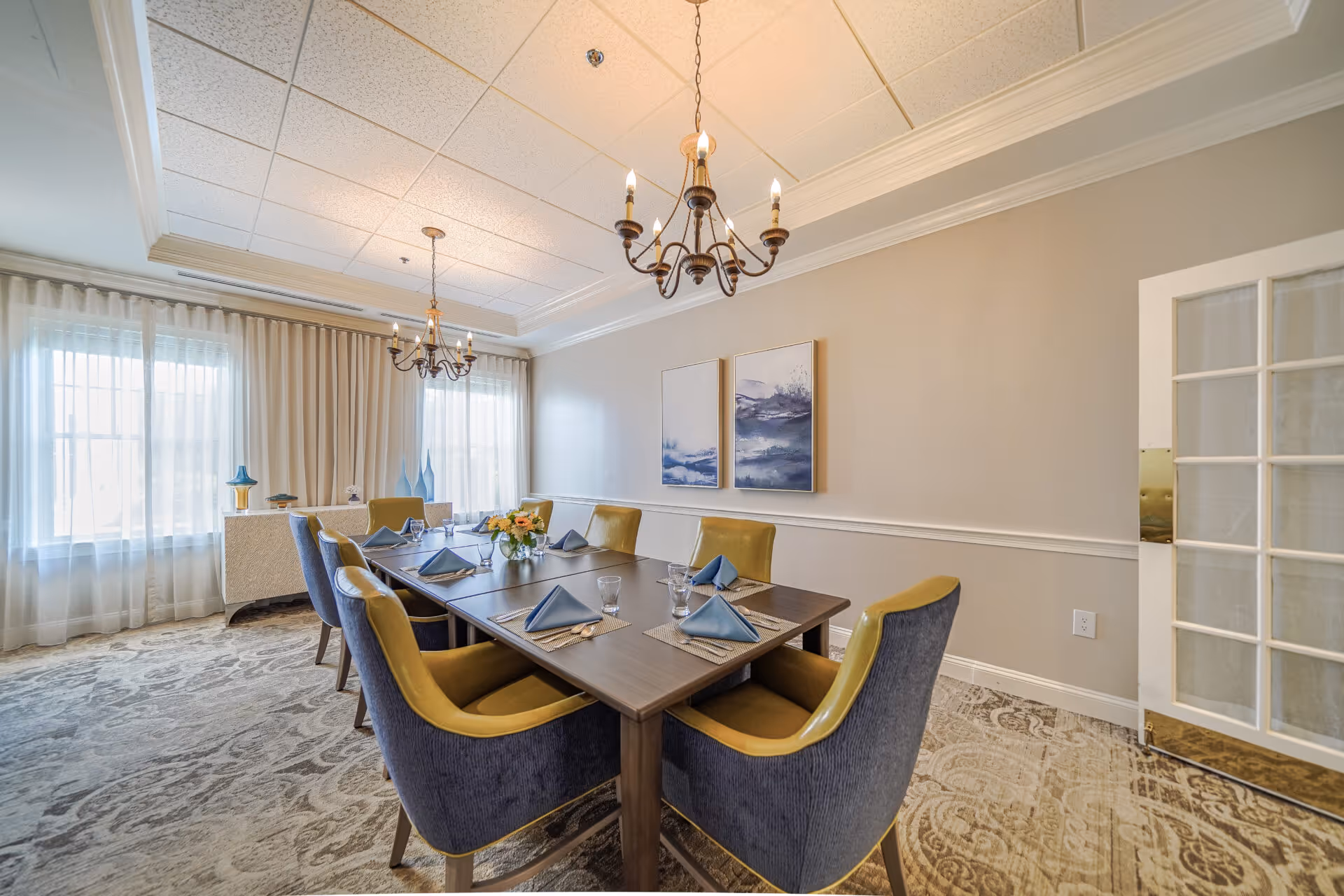 Well-lit dining room with a rectangular table set for six, upholstered chairs, chandeliers, and large windows with curtains.