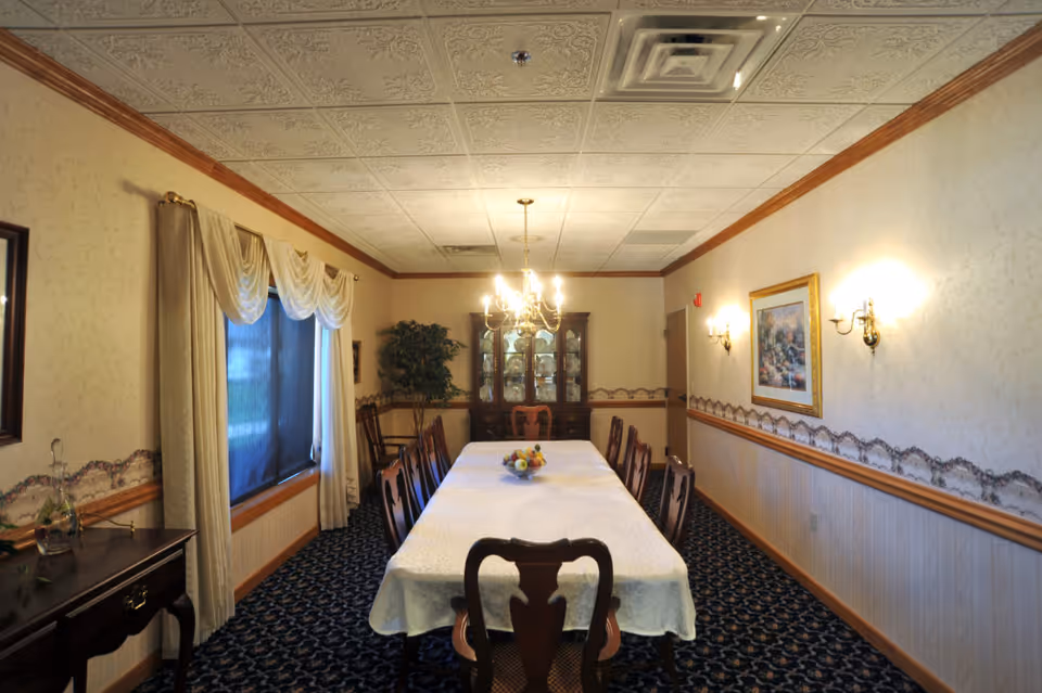 A formal dining room with a long rectangular table covered with a white tablecloth, surrounded by wooden chairs. A chandelier hangs above the table, and a bowl of fruit is placed in the center. The room has patterned carpet, wallpaper with a decorative border, wall sconces, a window with curtains, a side table, a framed painting, and a glass-front cabinet filled with dishes.
