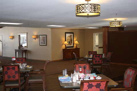 Dining room with tables set for meals, red-patterned chairs, decorative overhead lights, and a sideboard with framed art.