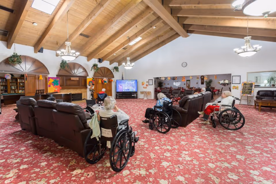 A spacious senior living facility common room with a high wooden beam ceiling and floral carpet. Several elderly residents in wheelchairs are seated facing a large flat-screen TV mounted on the far wall. The room features brown leather sofas arranged in rows, a fireplace with wooden paneling, hanging plants, and colorful decorations. A large mirror on the right wall reflects a dining area with tables and chairs.