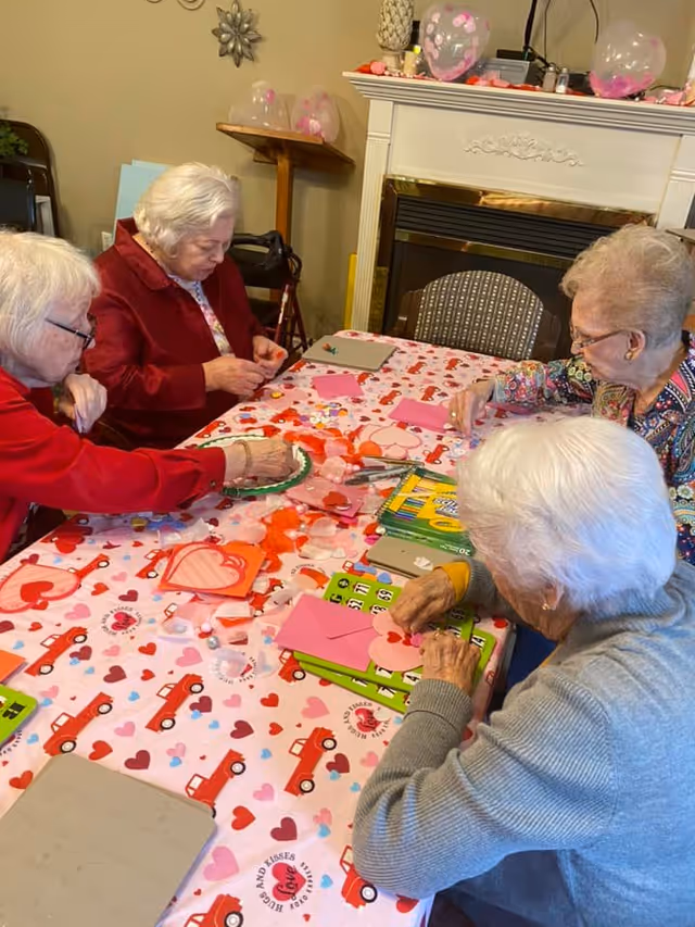 Four elderly women sitting around a table covered with a pink Valentine-themed tablecloth, engaged in crafting activities with paper hearts, envelopes, and other decorative materials. The room has a fireplace in the background with pink balloons and decorations on the mantle.