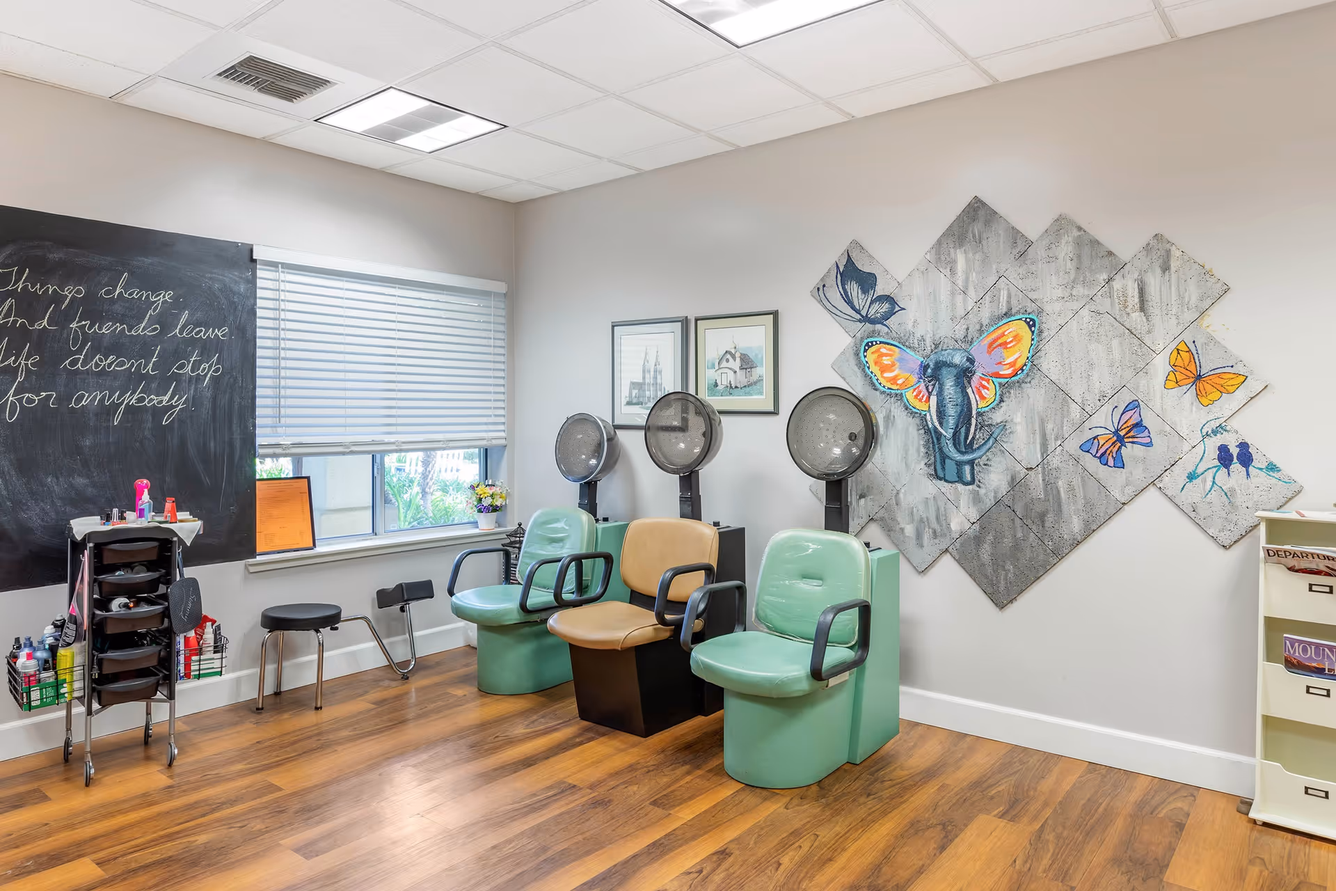 A salon room with three seated hair dryers, colorful elephant and butterfly wall art, a chalkboard, and salon supplies by a window.