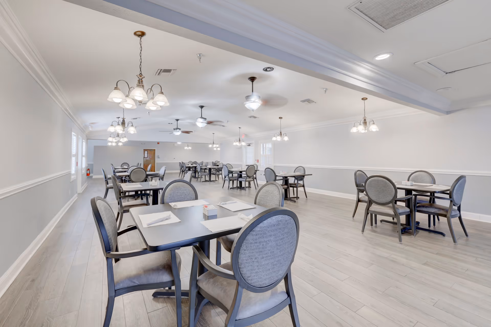 A spacious dining room with multiple tables and chairs arranged neatly. The room has light-colored walls with a chair rail, light wood flooring, ceiling fans, and hanging light fixtures. Each table is set with placemats, napkins, and a small condiment holder.