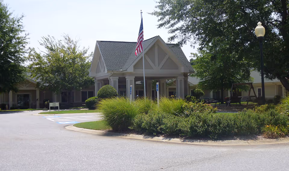Front entrance of a senior living facility with a covered porte-cochère, an American flag, landscaped circular driveway and benches.
