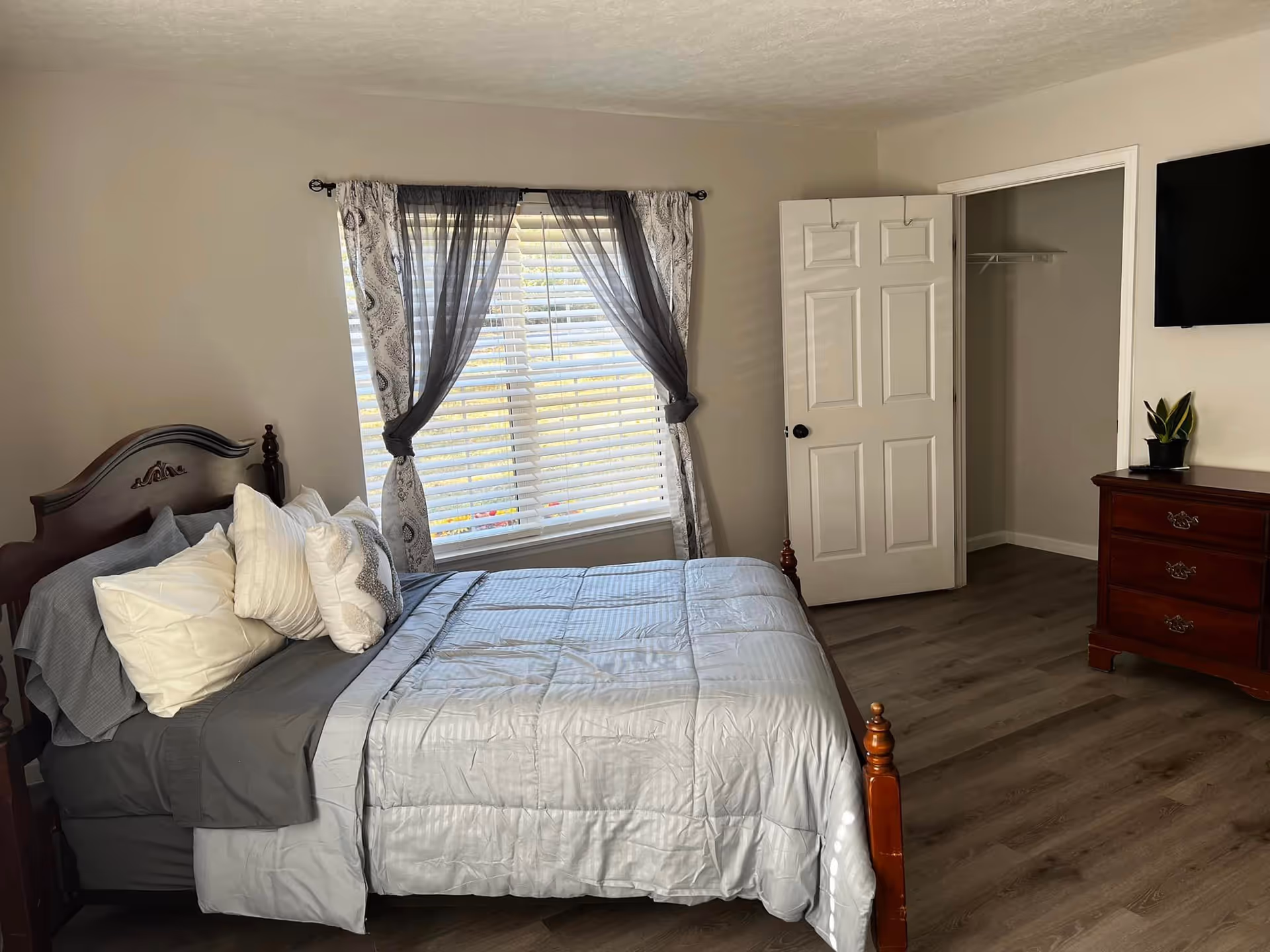 A neatly made bed with multiple pillows in a bedroom featuring a wooden headboard. There is a window with sheer and patterned curtains letting in natural light. An open door reveals a closet with a hanging rod. A wooden dresser with a small potted plant on top is positioned below a wall-mounted flat screen TV. The room has light-colored walls and wood flooring.