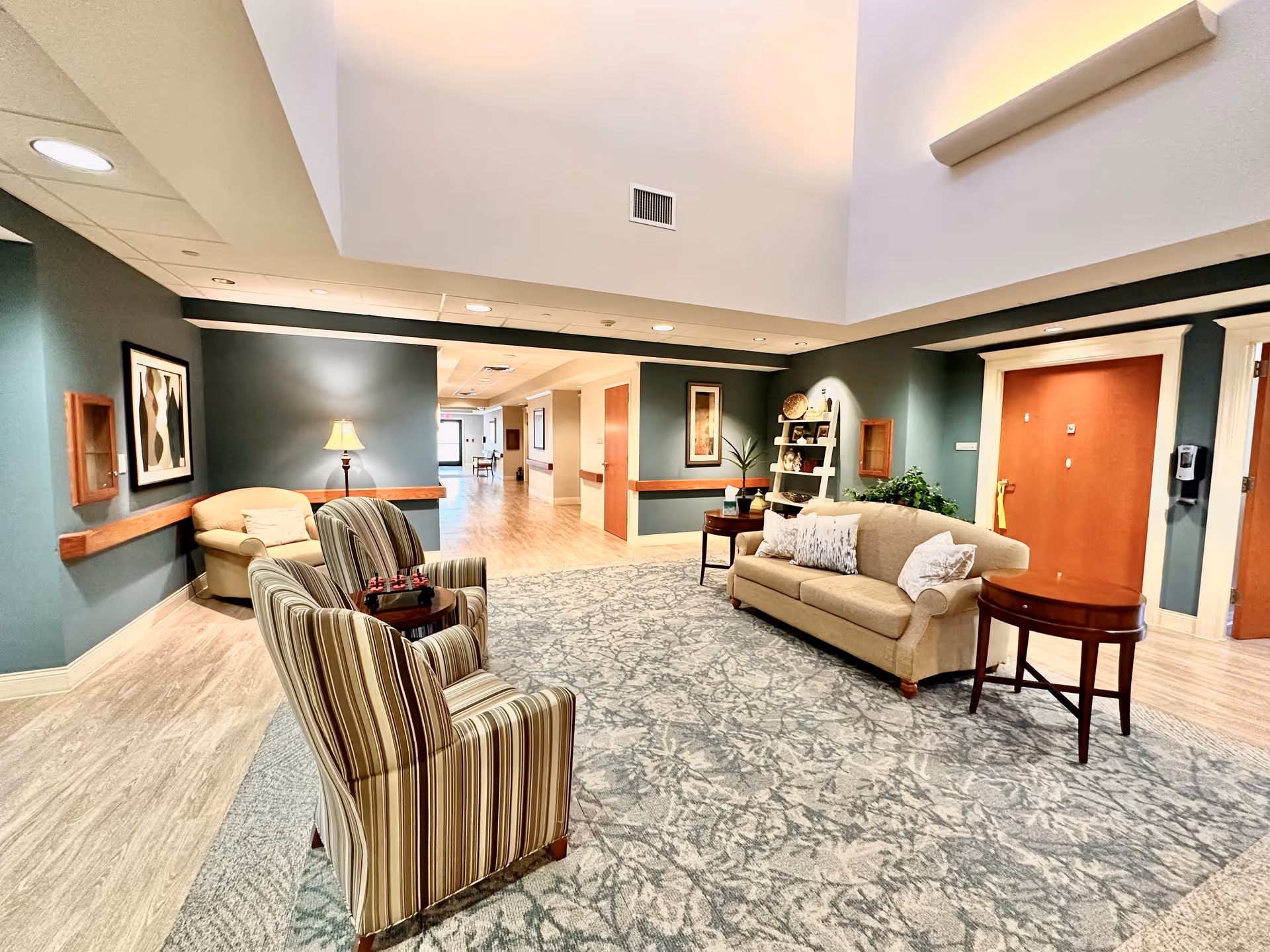 A cozy sitting area in a senior living facility with a beige sofa, two striped armchairs, a round wooden side table, and a lamp. The walls are painted green with framed artwork and decorative shelves. The floor is covered with a patterned carpet and light wood flooring. There is a hallway leading to other rooms in the background.
