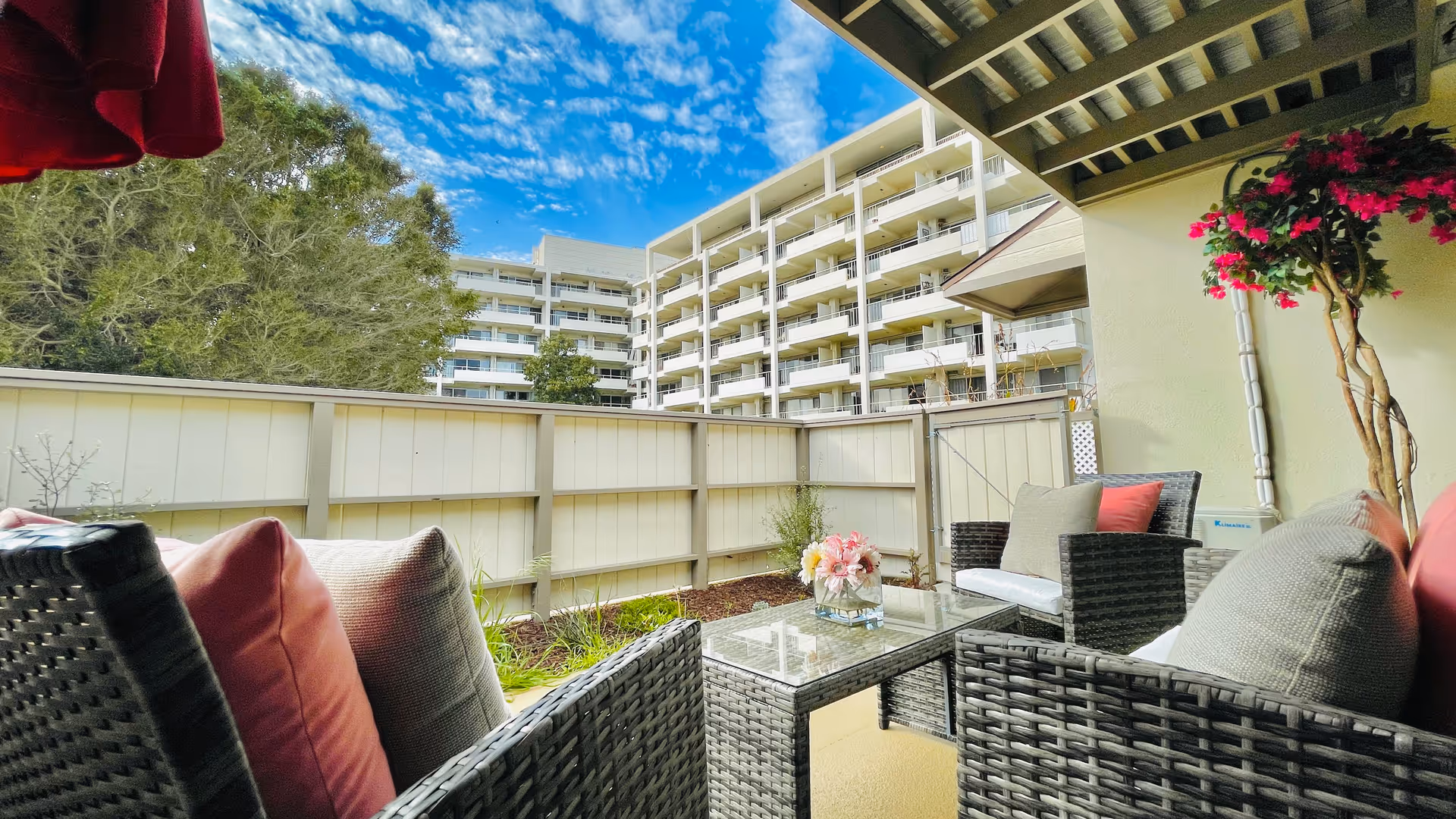Outdoor patio area with wicker furniture including chairs and a glass-top table adorned with a flower arrangement. The patio is enclosed by a white fence and overlooks a multi-story residential building under a blue sky with scattered clouds. There are plants and a tree visible around the patio.