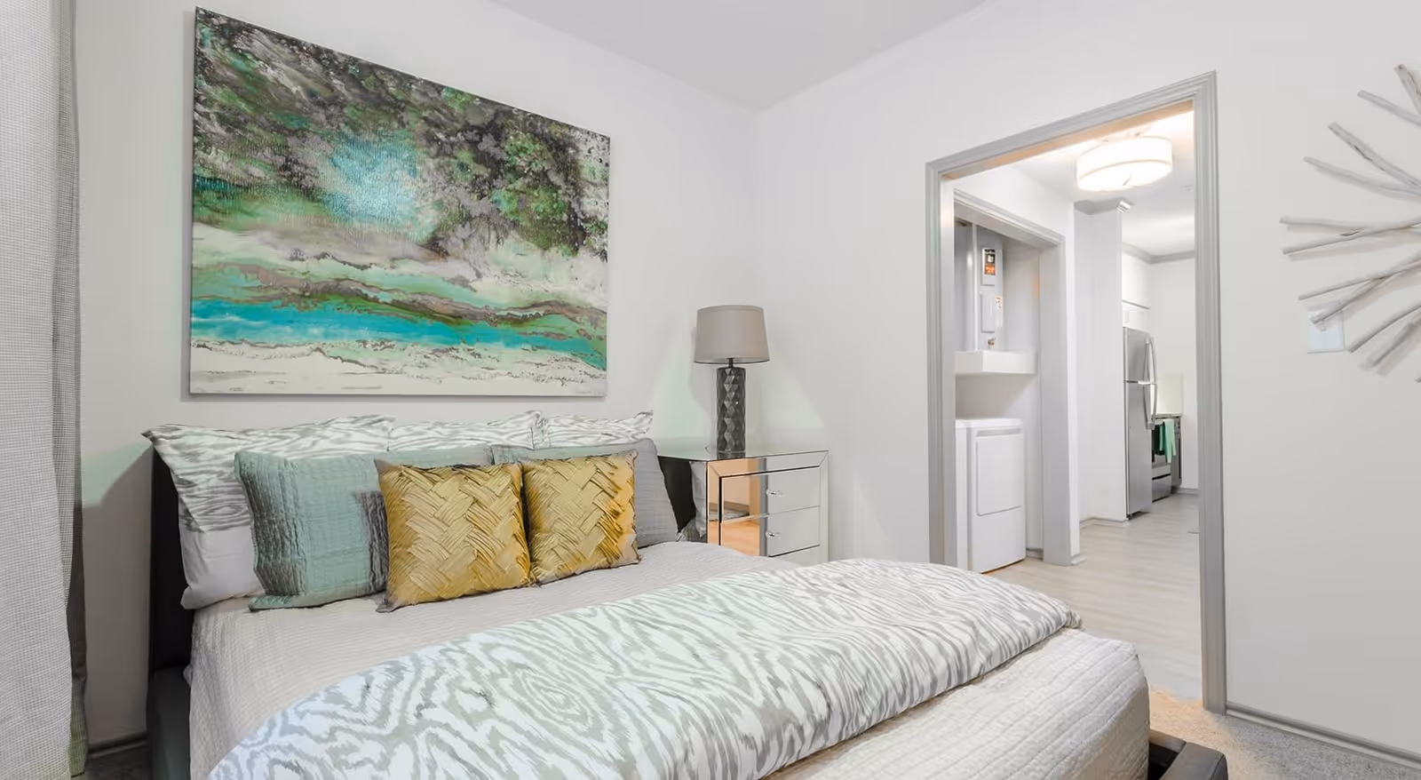 A modern bedroom with a neatly made bed featuring patterned bedding and multiple pillows in shades of white, green, and yellow. Above the bed hangs a large abstract painting with green, white, and black colors. To the right of the bed is a mirrored nightstand with a lamp. The room opens into a hallway leading to a kitchen area with visible appliances and a laundry area with a stacked washer and dryer.