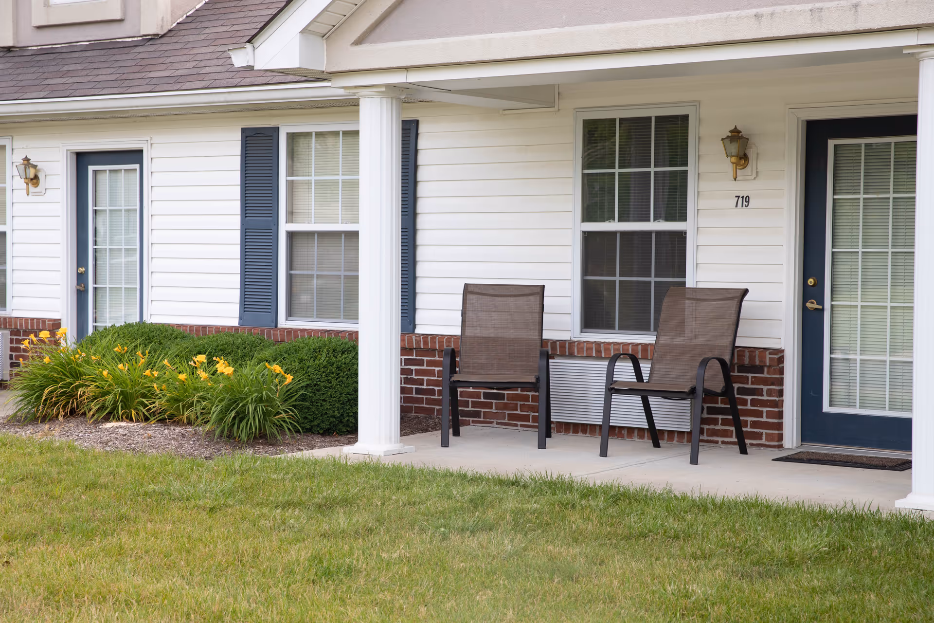 Outdoor patio area of Meadow Lakes Assisted Living and Garden Homes featuring two brown mesh chairs on a concrete porch, white siding with blue shutters, two windows, a blue door with glass panes, a white column, and a garden bed with yellow flowers and green shrubs.