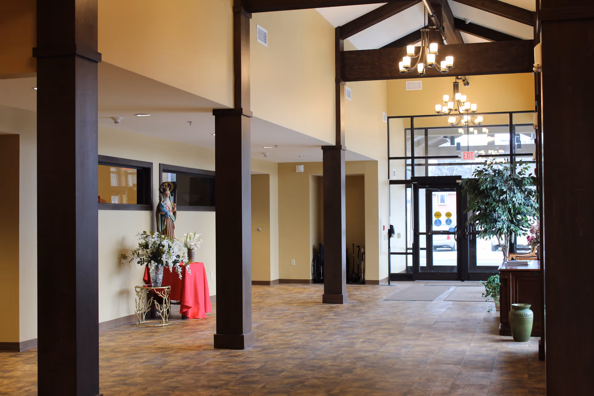 Spacious senior living facility lobby with high ceilings, wooden beams, and chandeliers. There are large glass doors at the entrance, a statue on a red-covered table with floral arrangements, and a potted plant near a wooden cabinet.