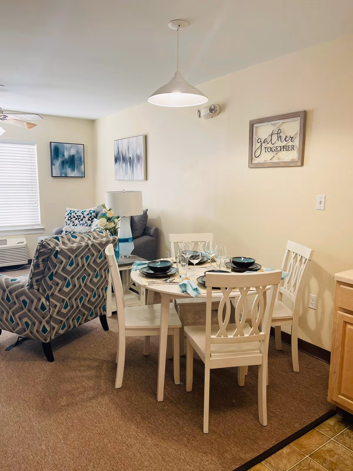 A cozy dining and living area in a senior living community. The dining area features a round white table set with four chairs, black plates, wine glasses, and teal napkins. Behind the dining area is a living space with a patterned armchair, a gray sofa, a white table lamp, and two abstract paintings on the wall. A framed sign on the wall reads 'gather TOGETHER'. The room has beige walls and carpeted flooring.