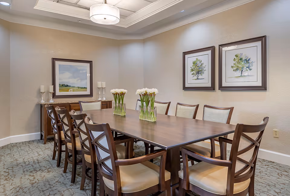 A dining room with a long wooden table surrounded by ten cushioned chairs. Three vases with white flowers are placed on the table. The walls are decorated with three framed paintings of trees and a landscape. A wooden sideboard with candles is against the wall, and a ceiling light fixture hangs above the table.