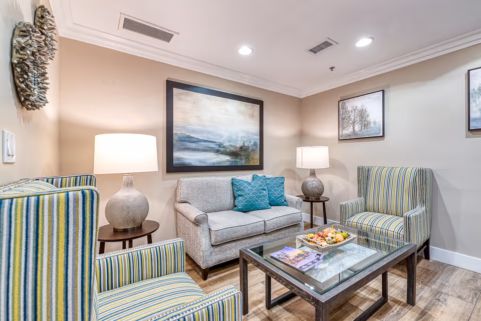 A cozy senior living facility lounge area with a gray sofa adorned with two blue patterned pillows, two striped armchairs, a glass coffee table with decorative items and brochures, two side tables each holding a lamp, and framed landscape artwork on beige walls.