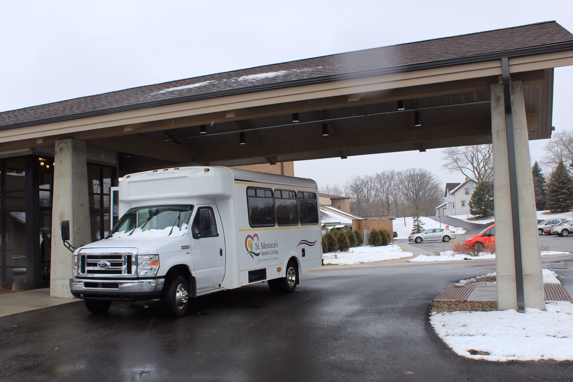 A St. Monica's Senior Living shuttle bus parked under the facility's covered entrance with snow on the ground and nearby buildings.