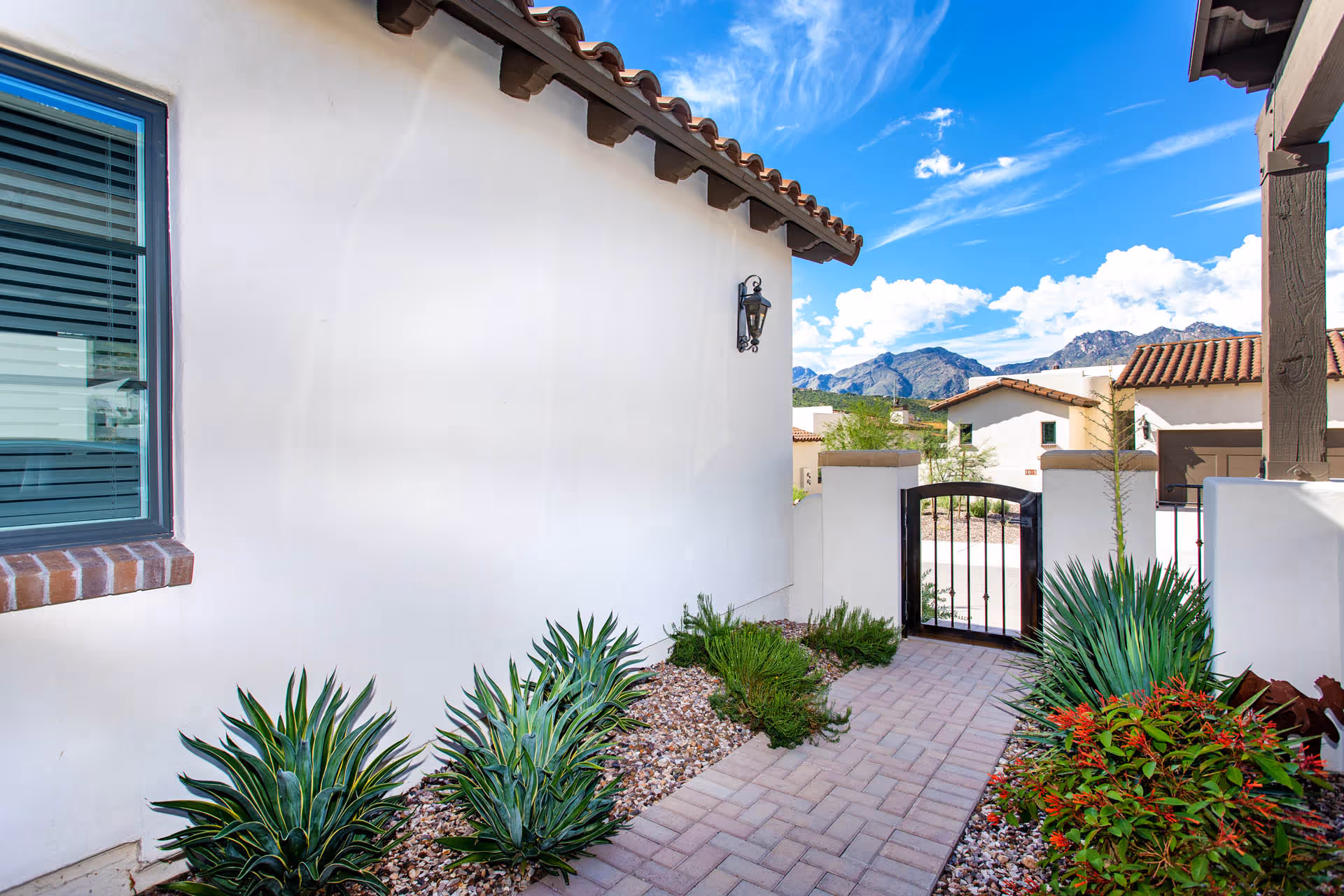 Outdoor pathway alongside a white stucco building with a window and a black metal gate at the end. The pathway is lined with green plants and small rocks, with a clear blue sky and mountains visible in the background.