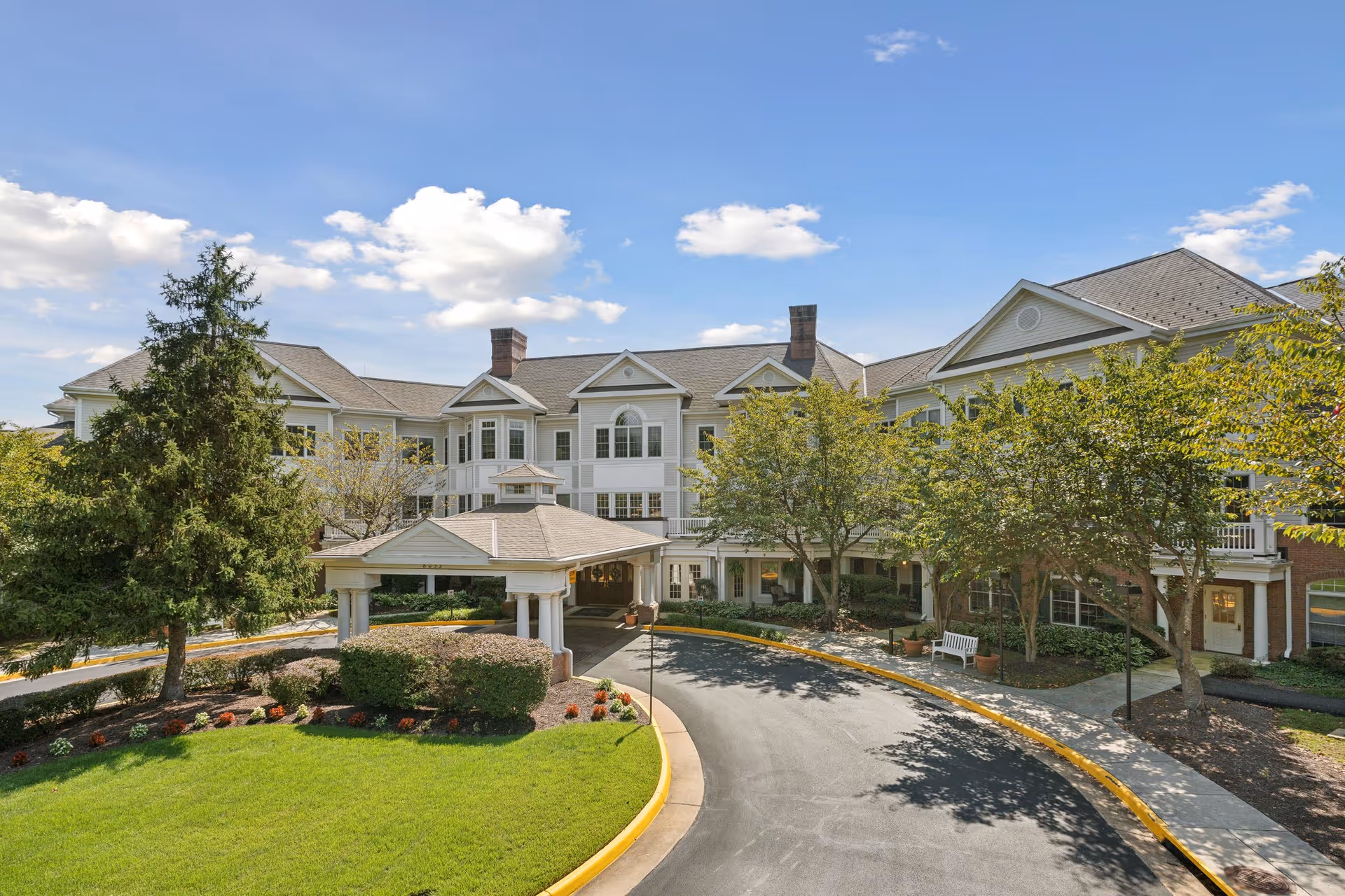 Front exterior of a multi-story senior living building with a covered circular driveway, landscaped lawn, and trees under a blue sky.
