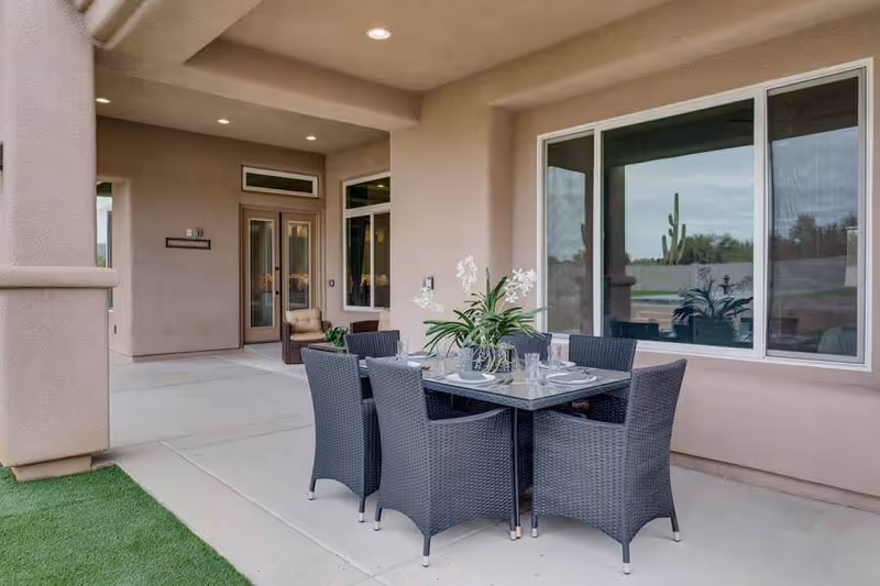 Outdoor covered patio area with a dining table set for six, featuring black wicker chairs and a glass-top table with a floral centerpiece. The patio is adjacent to a building with large windows and a door, and there is artificial grass visible on the left side.