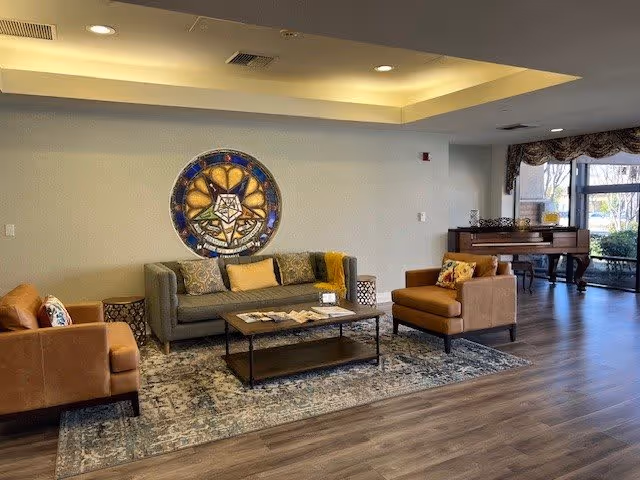A cozy living room area in a senior living community featuring a gray sofa with decorative pillows, two brown armchairs, a coffee table with magazines, a patterned rug, and a large decorative circular wall art. In the background, there is a piano near large windows with curtains allowing natural light to enter.