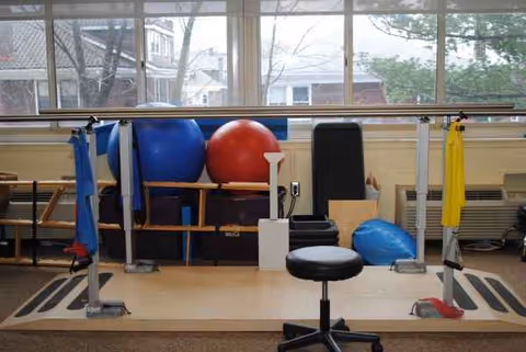 Rehabilitation gym area with parallel bars, exercise balls, and a chair by large windows.