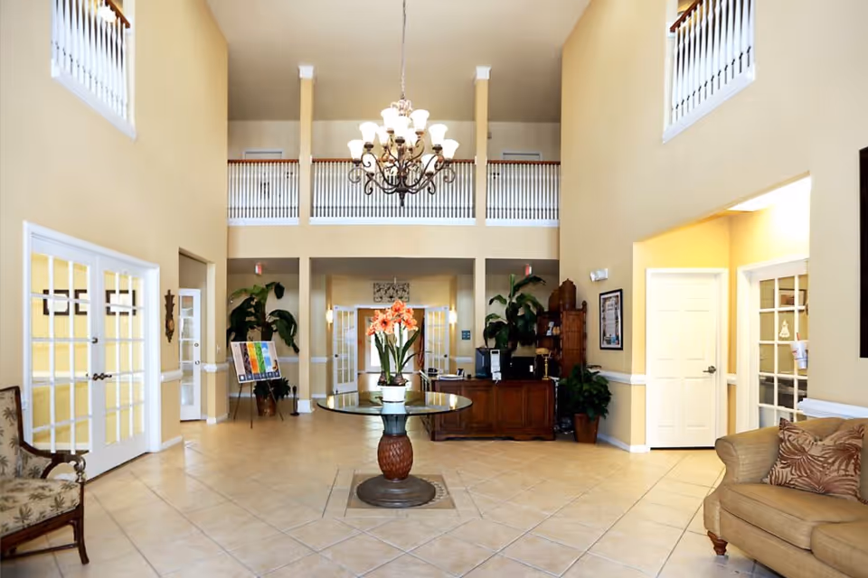 Bright two-story senior living lobby with a central round table and flowers, chandelier, reception desk, seating, and potted plants.