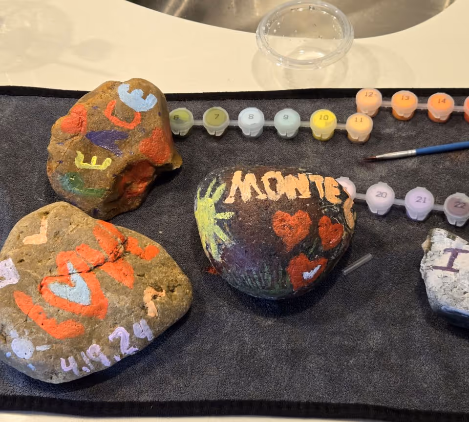 Painted river rocks decorated with hearts and names sit on a towel next to numbered paint pots and a paintbrush.