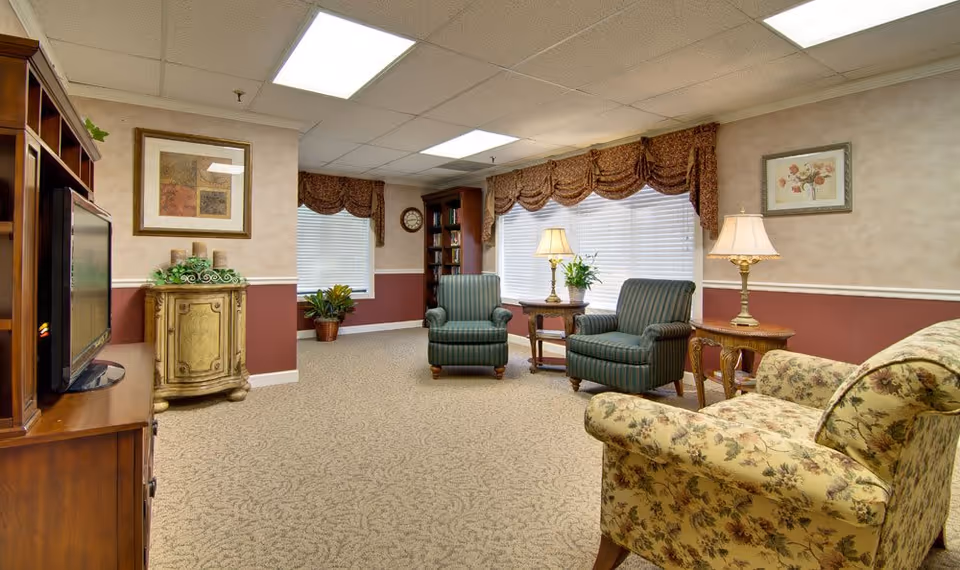 A cozy living room with patterned carpet, two striped armchairs, a floral upholstered sofa, wooden side tables with lamps, a wooden entertainment center with a TV, framed artwork on the walls, and large windows with blinds and decorative valances.