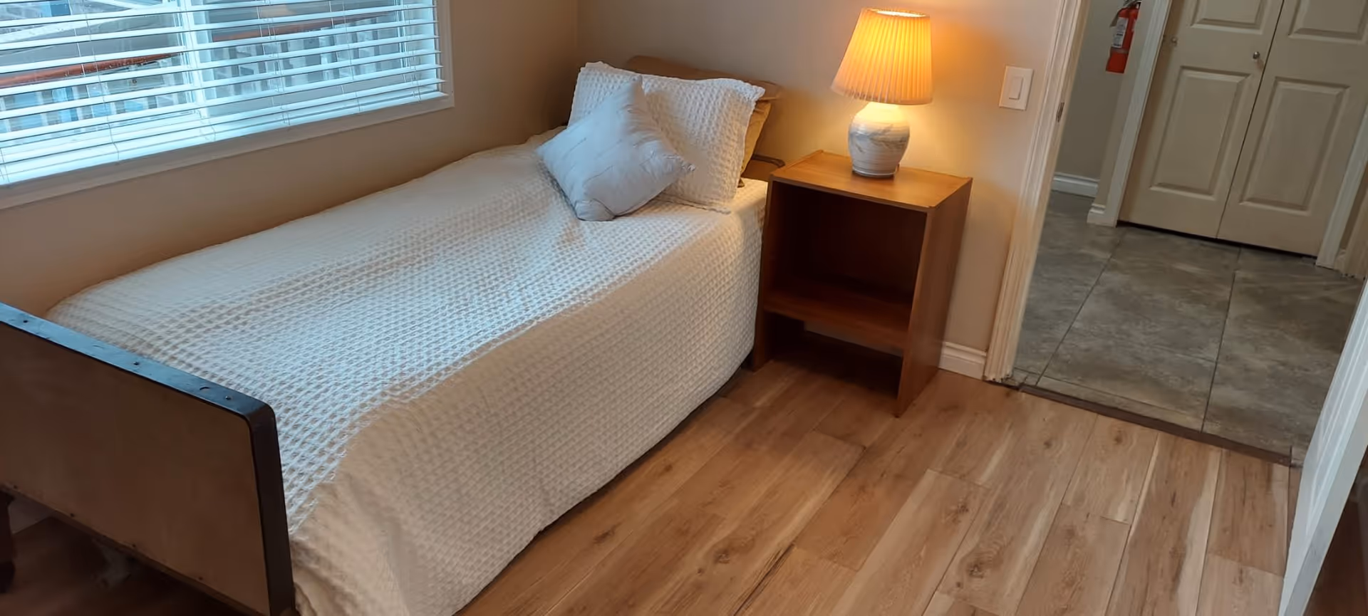 A small bedroom with a single bed covered in a white textured bedspread and two pillows. Next to the bed is a wooden nightstand with a lit table lamp. The room has wooden flooring and a window with white blinds. A doorway leads to a tiled floor area with a closed closet door and a fire extinguisher mounted on the wall.