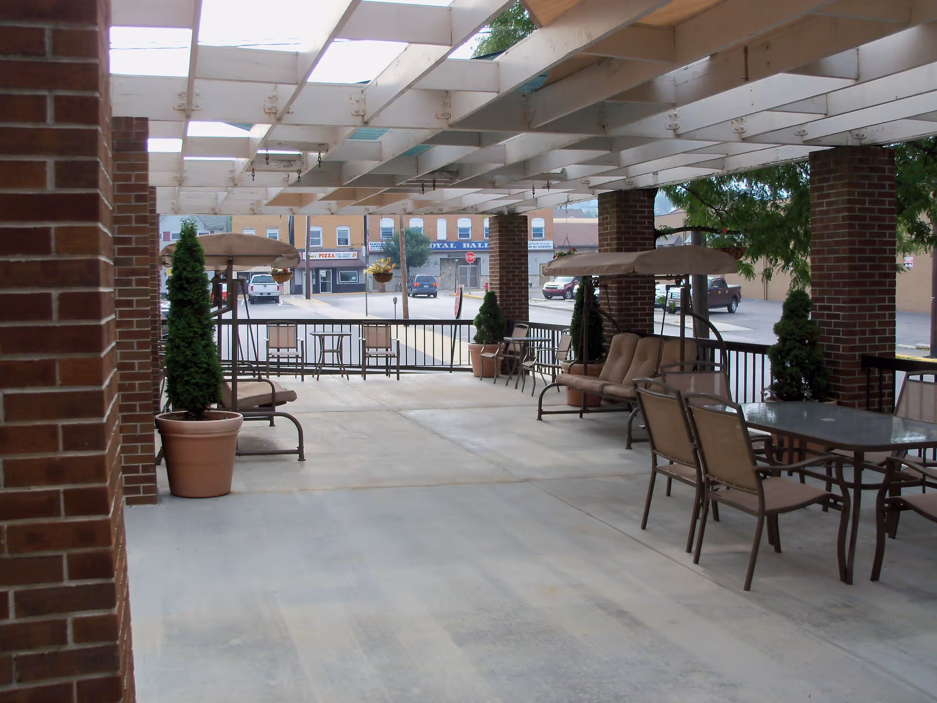 Covered outdoor patio area with brick pillars, potted plants, cushioned swing benches with canopies, and a glass-top table with chairs. The patio overlooks a street with parked cars and storefronts in the background.