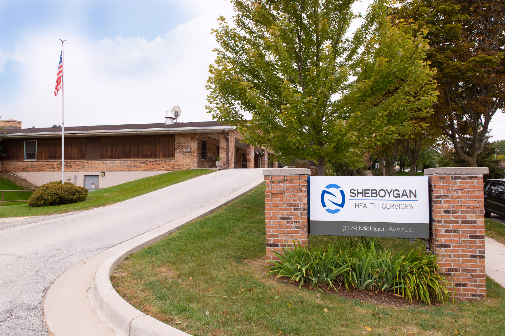 Exterior view of Sheboygan Health Services building with a brick sign displaying the facility name and address at 3129 Michigan Avenue. The building is a single-story brick structure with a driveway leading up to the entrance. There is an American flag on a flagpole to the left and trees surrounding the area.