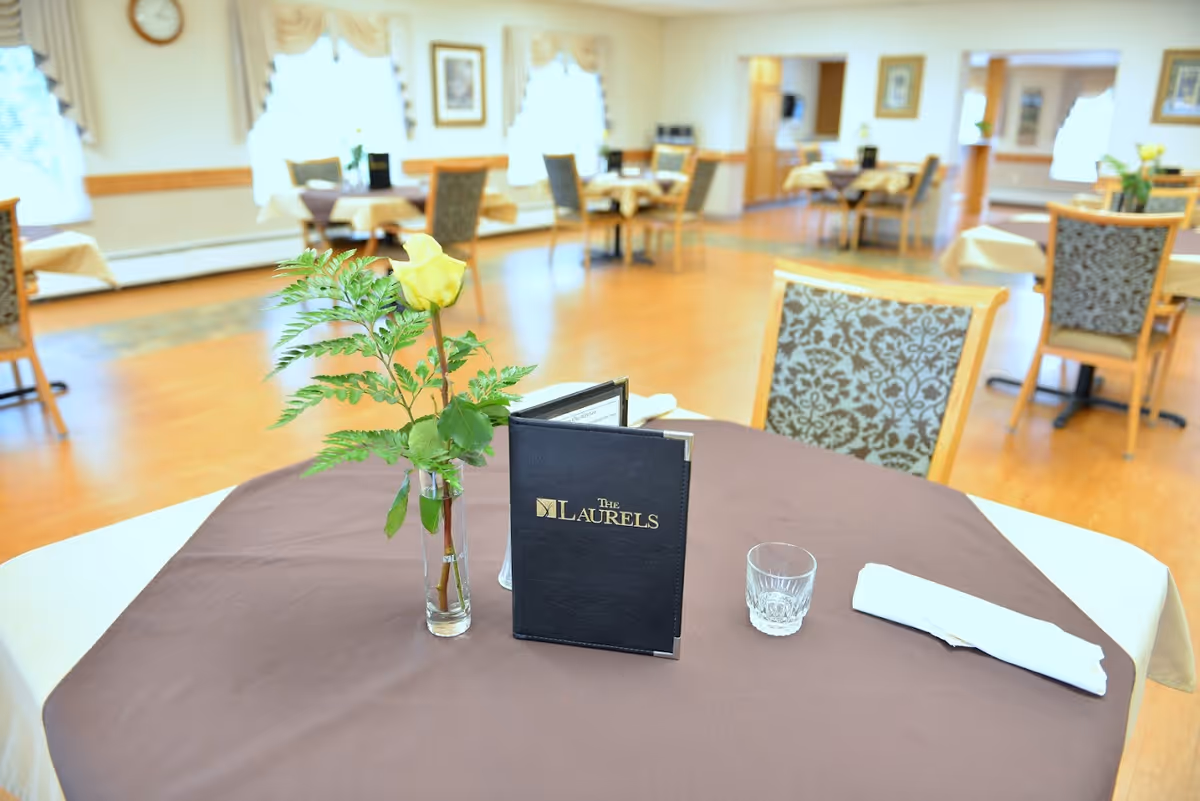 A dining room in a senior living facility with tables covered in brown and beige tablecloths. One table in the foreground has a small vase with a yellow rose and greenery, a glass, a folded white napkin, and a black menu folder labeled 'The Laurels'. The room has wooden floors, patterned chairs, and windows with sheer curtains letting in natural light.