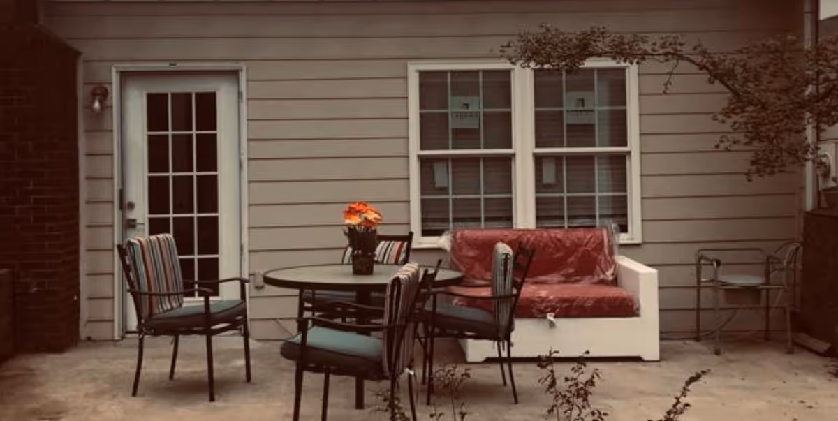 Outdoor patio area with a round table surrounded by four chairs with striped cushions, a white bench covered with a red protective cover, a potted plant with orange flowers on the table, and a glass door and two windows on the beige siding wall of the building.