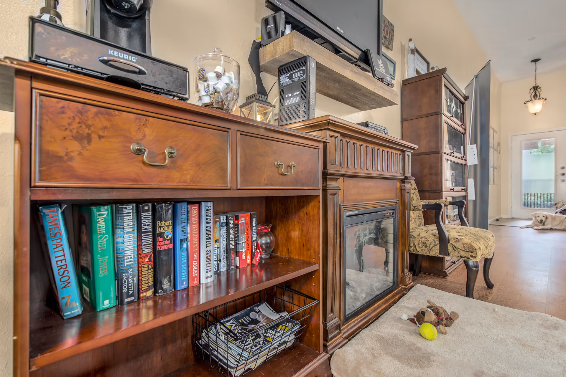 Interior view of a cozy living room area featuring a wooden cabinet with books and magazines, a Keurig coffee maker on top, a fireplace, an upholstered armchair, and a dog lying on the floor near the entrance door in the background.