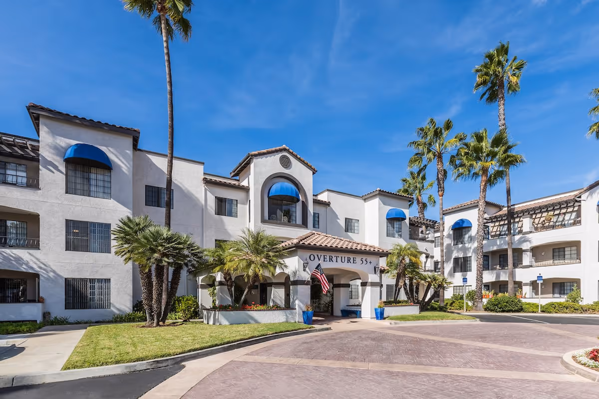 Exterior view of Overture San Marcos senior living facility with white buildings, blue window awnings, palm trees, and a clear blue sky.