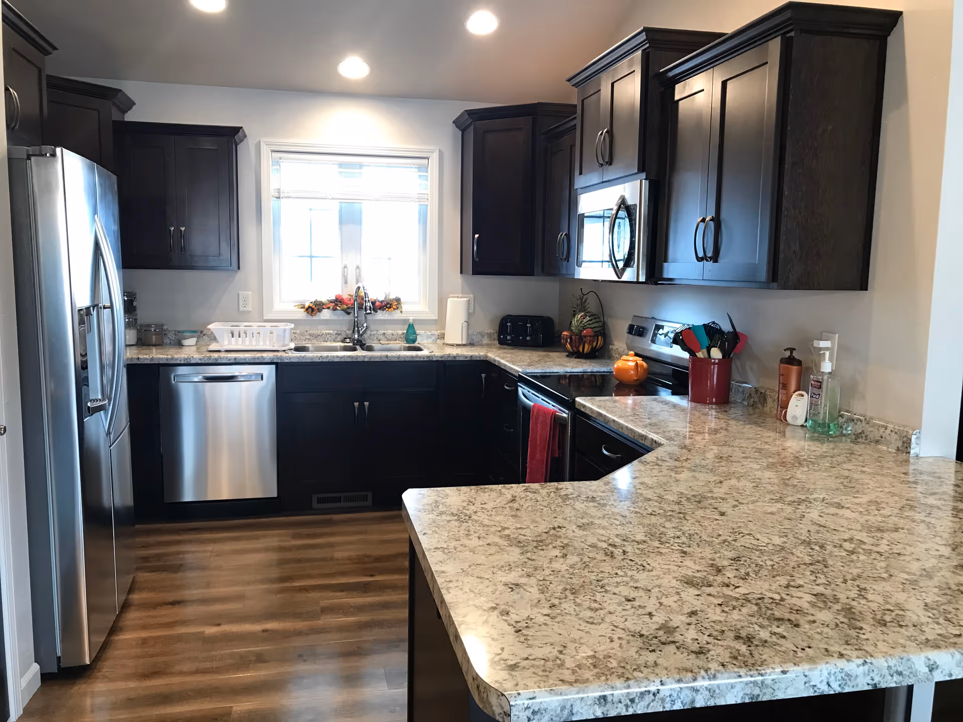 Modern kitchen with dark wood cabinets, stainless steel appliances, and a large speckled countertop bar under recessed lighting.