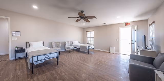 A spacious room with two single beds, two chairs, a ceiling fan, a TV on a stand, and a gray armchair. The room has wooden flooring, white walls, and windows letting in natural light. An exit door is open, showing bright light outside.