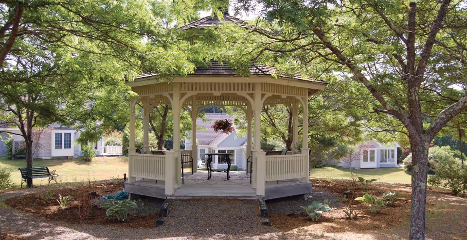 A white wooden gazebo with a shingled roof situated in a garden area surrounded by trees and plants. There is a black metal table and chairs inside the gazebo, and a hanging flower basket is visible. In the background, there are residential buildings with large windows and well-maintained lawns.