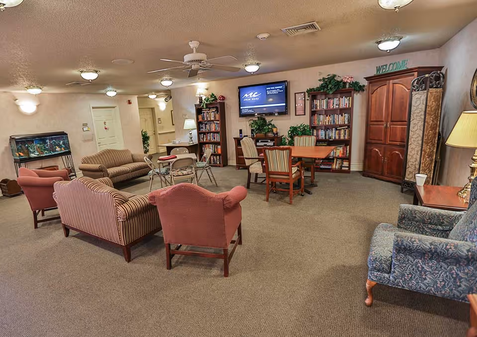 A cozy common room in a senior living facility with various seating options including striped and patterned armchairs and sofas arranged around small tables. There is a large flat-screen TV mounted on the wall above bookshelves filled with books and decorative plants. A fish tank is visible on the left side of the room. The room has carpeted floors, ceiling fans, and soft lighting fixtures. A wooden cabinet and a lamp are on the right side, and a 'WELCOME' sign is displayed above the bookshelf.