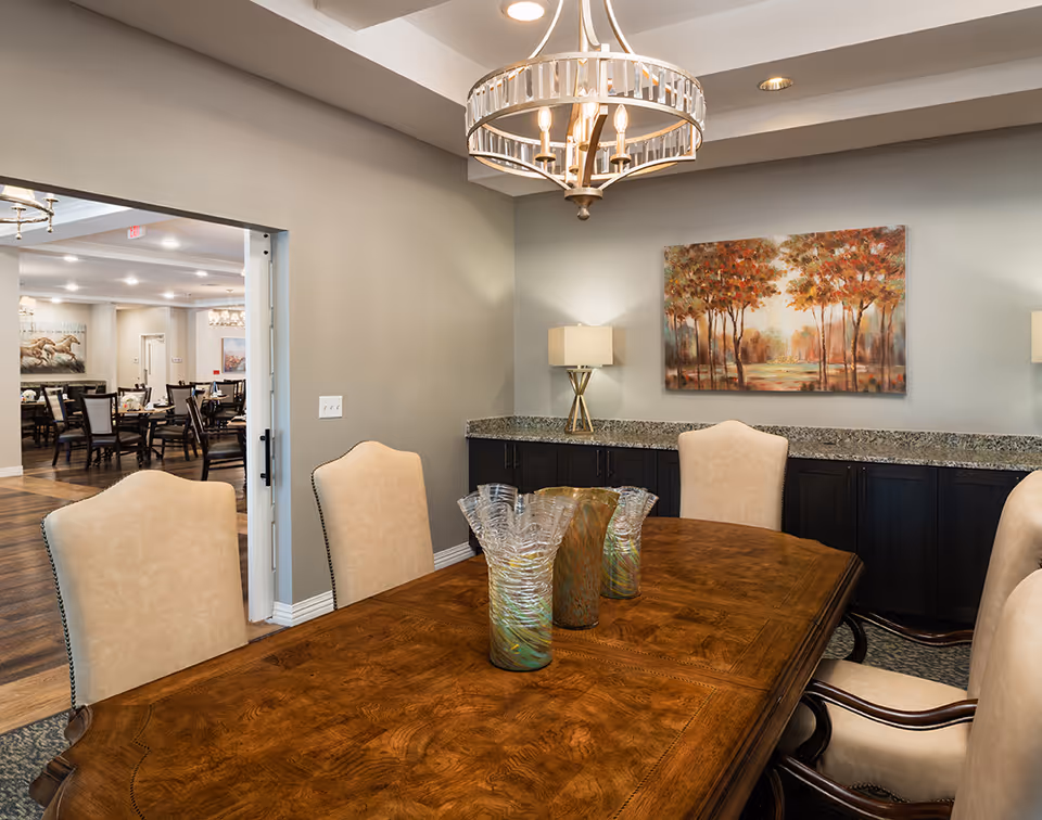 Elegant dining room with a polished wooden table, upholstered chairs, a chandelier, and a sideboard topped by a landscape painting.