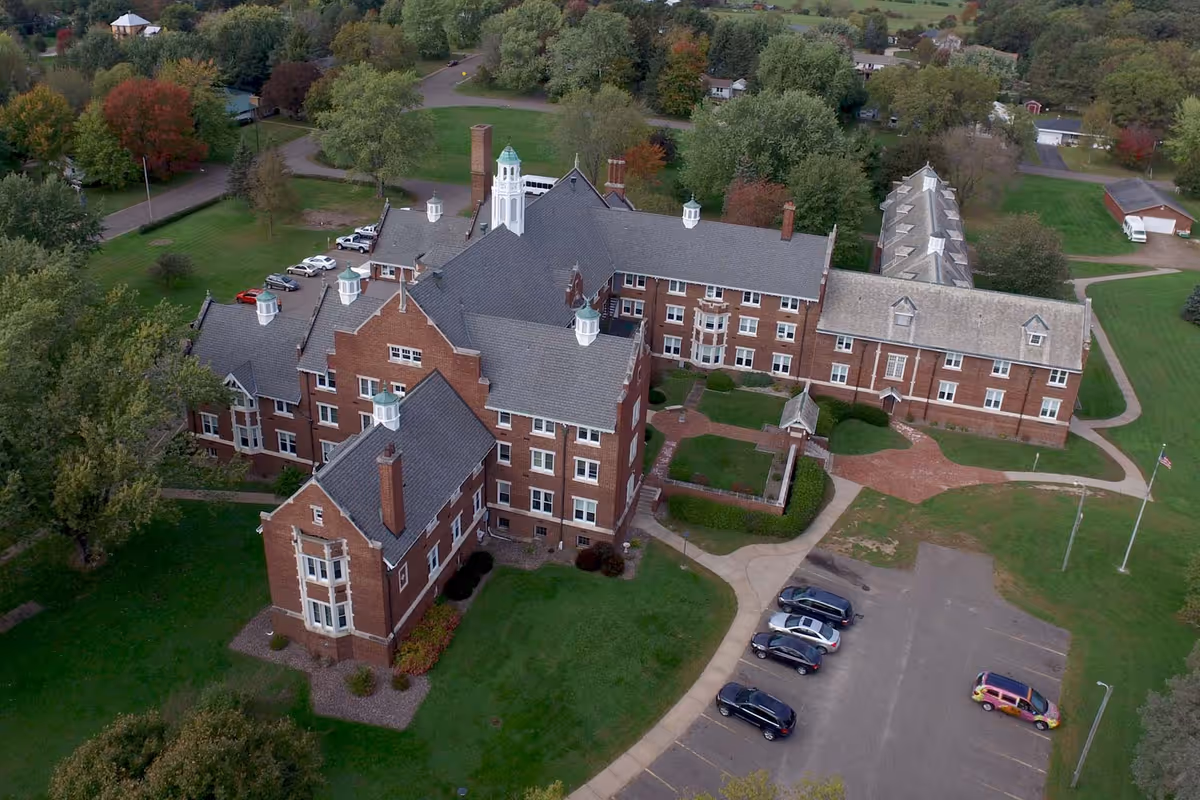 Aerial view of a large brick building complex with multiple wings and white cupolas on the roofs, surrounded by green lawns, trees, and a parking lot with several cars. The setting appears to be a senior living facility with pathways and landscaped areas.