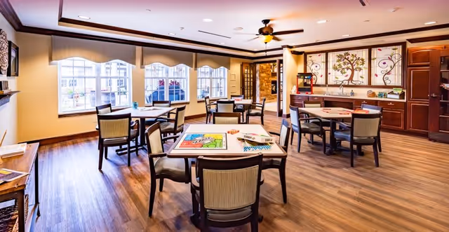 Well-lit communal dining/activity room with multiple square tables and chairs, large windows, hardwood floors, and a cabinet counter area.