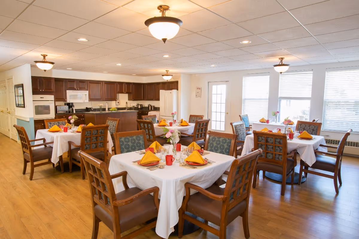 A bright dining room with several tables covered in white tablecloths, each set with yellow folded napkins, red mugs, glassware, and floral centerpieces. The room features wooden chairs with patterned cushions, hardwood floors, and a kitchen area with dark wooden cabinets, a white refrigerator, and a microwave in the background. Large windows with blinds allow natural light to fill the space.