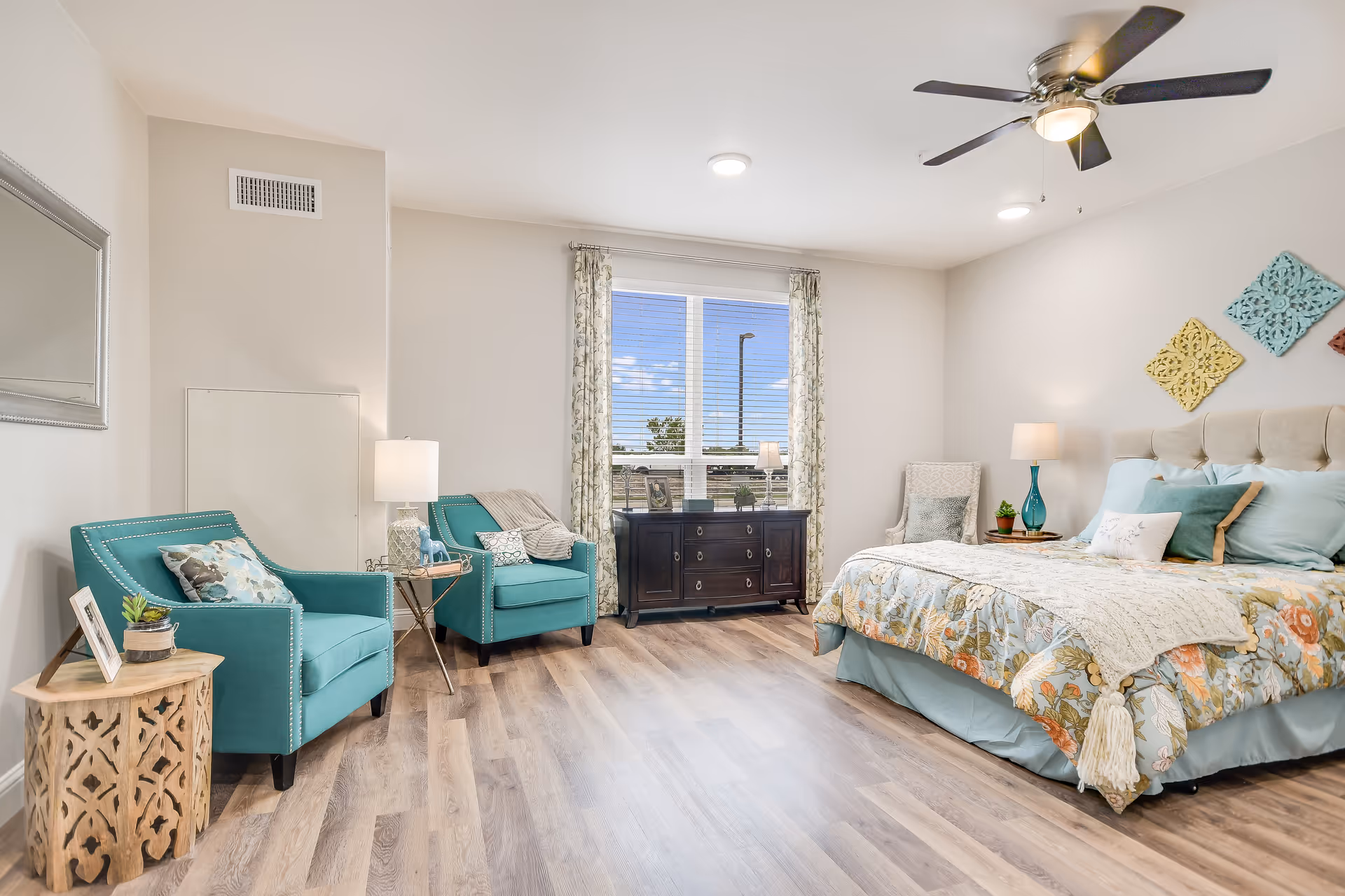 A bright and spacious bedroom in a senior living facility featuring a large bed with floral bedding and multiple pillows, two teal armchairs with decorative pillows, a wooden side table with a small plant and framed photo, a dark wooden dresser under a window with floral curtains, and a ceiling fan with lights.