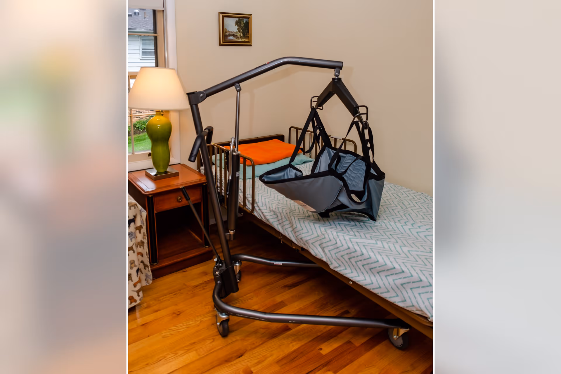A bedroom in an assisted living facility featuring a hospital-style bed with a patterned bed sheet and an orange blanket. Next to the bed is a wooden nightstand with a green lamp. A patient lift device with a sling is positioned over the bed. A window and a framed picture are visible on the wall.