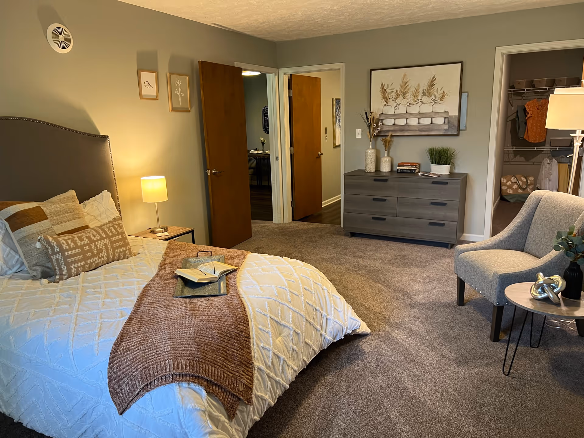 A cozy bedroom with a neatly made bed featuring white and brown bedding, a small bedside table with a lamp, a gray upholstered chair next to a small round table with decorative items, a dresser with vases and books, and an open closet with clothes and storage bins. Two wooden doors lead to other rooms.