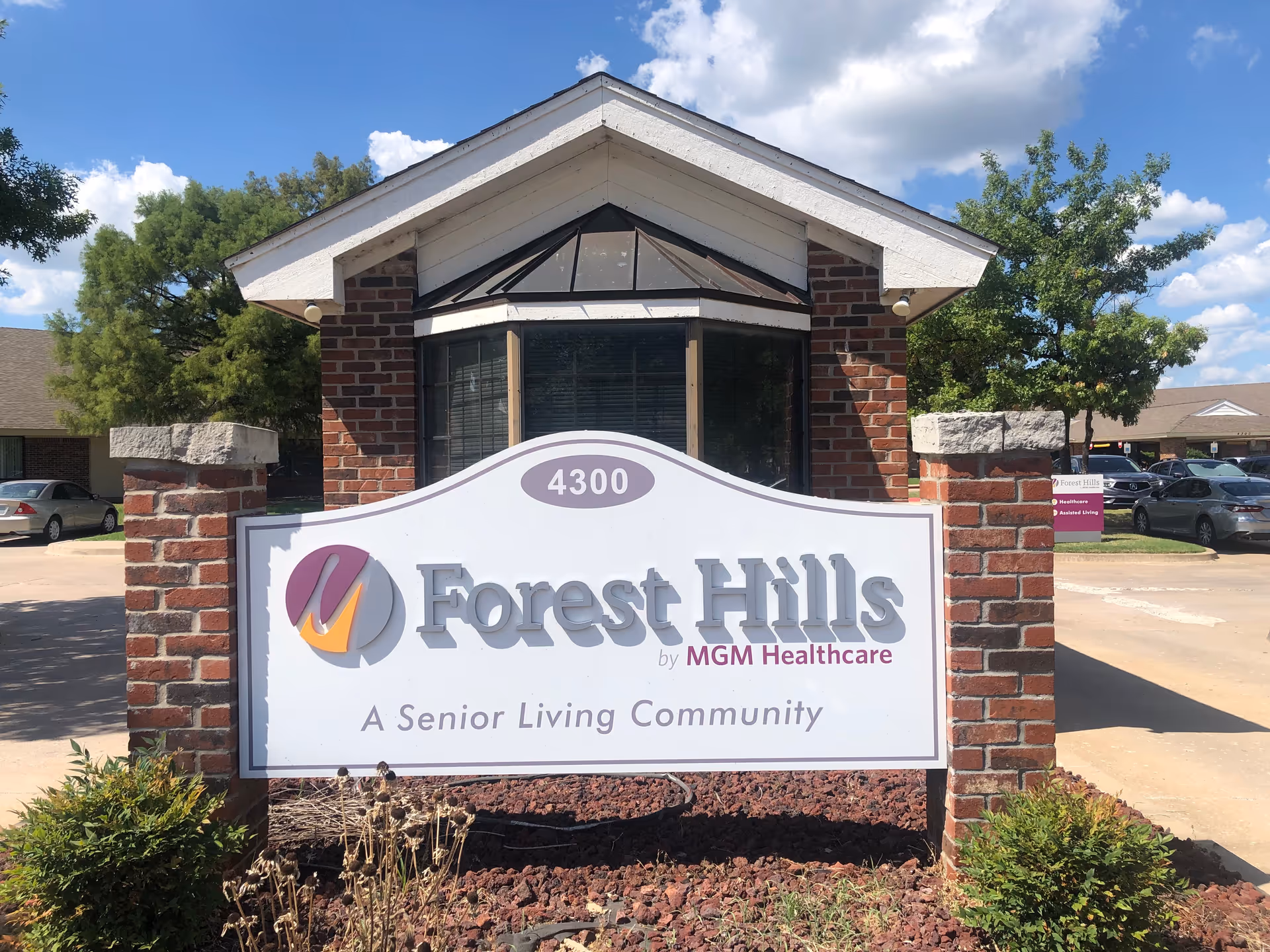 Outdoor view of the entrance sign for Forest Hills, a senior living community by MGM Healthcare, mounted on a brick structure with a small building and parked cars in the background under a partly cloudy sky.