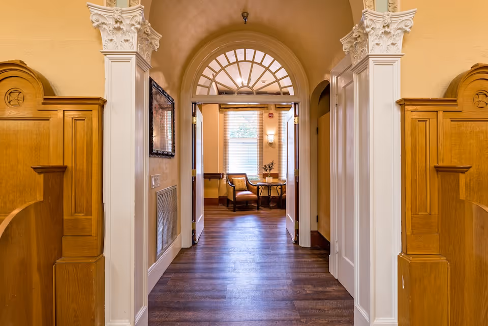 Ornate hallway with wooden paneling and an arched doorway leading to a small sitting area with chairs and a table.